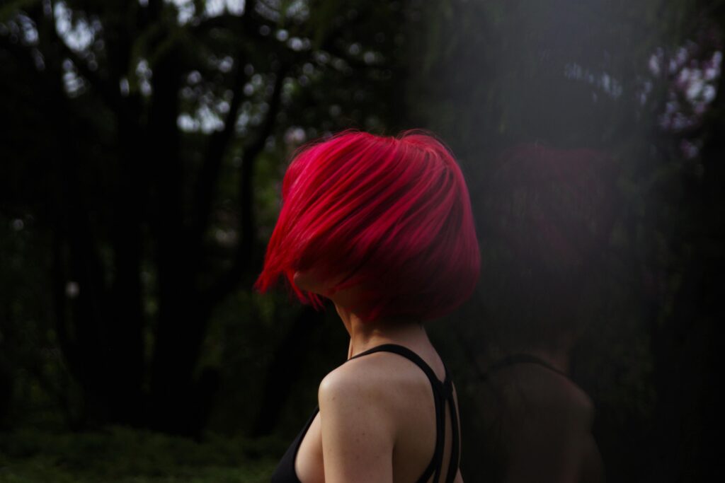 Woman with vibrant red hair turns away in a shadowy forest, striking dynamic pose in natural setting.
